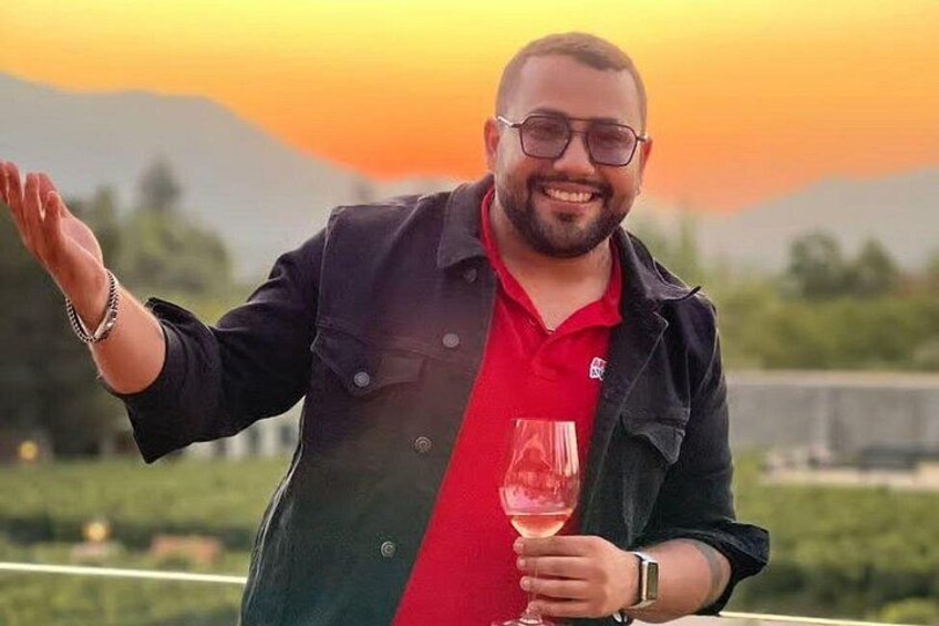 A person holding a glass of red wine, wearing a black jacket over a red shirt, standing outdoors with a sunset in the background. 2