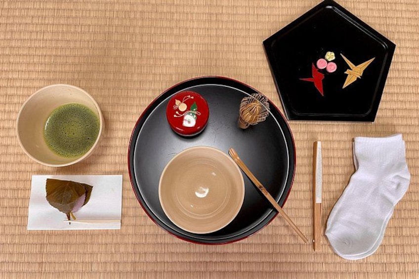 Preparación tradicional de una ceremonia del té japonesa, con una bandeja negra, una taza de té verde, una taza blanca, un cucharón rojo para té, un par de palillos, un paño blanco y una caja decorativa. 5