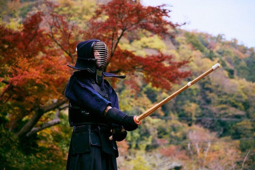 Une personne vêtue d’une tenue traditionnelle d’arts martiaux, tenant une arme, se tient dans une forêt où les arbres arborent les couleurs de l’automne. 4