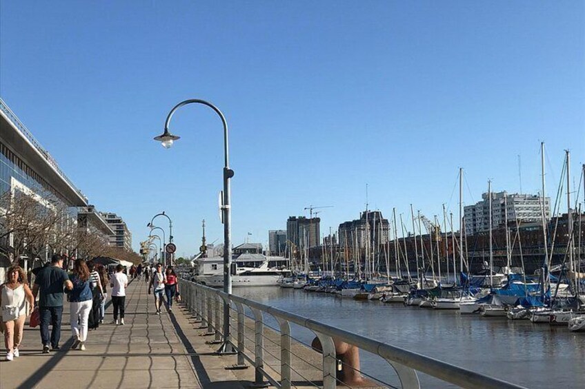 Un paseo marítimo con gente caminando, una hilera de barcos amarrados y edificios al fondo. 2