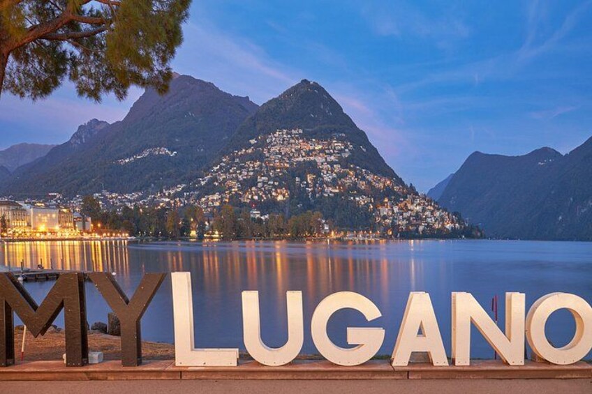 A lake with mountains in the background and the words 'MY LUGANO' in the foreground. 1