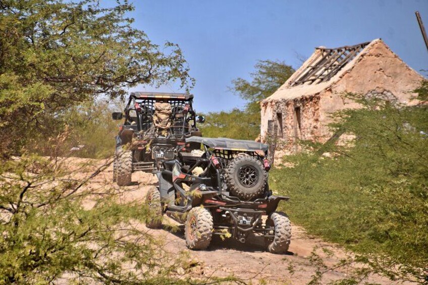 A 4x4 vehicle navigating a rocky terrain with sparse vegetation and a dilapidated building in the background. 3