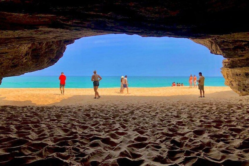 A beach scene with people walking and standing, a clear blue sky, and a rocky cave entrance. 4