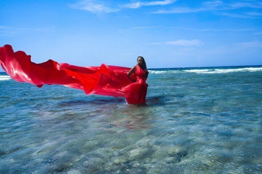 A person in a red dress standing in shallow water with waves in the background. 1