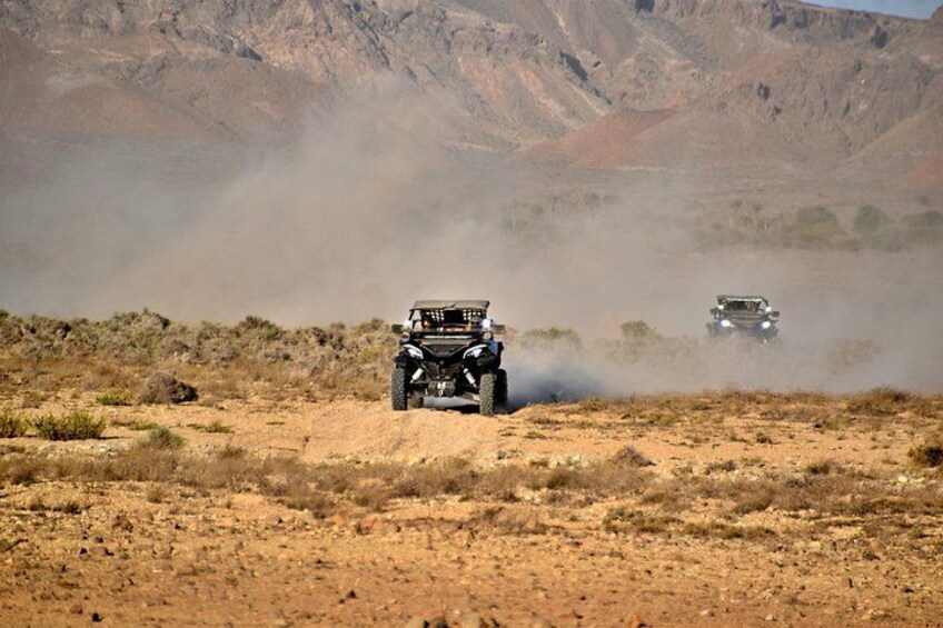 Two off-road vehicles driving on a dusty terrain with sparse vegetation and rocky hills in the background. 3