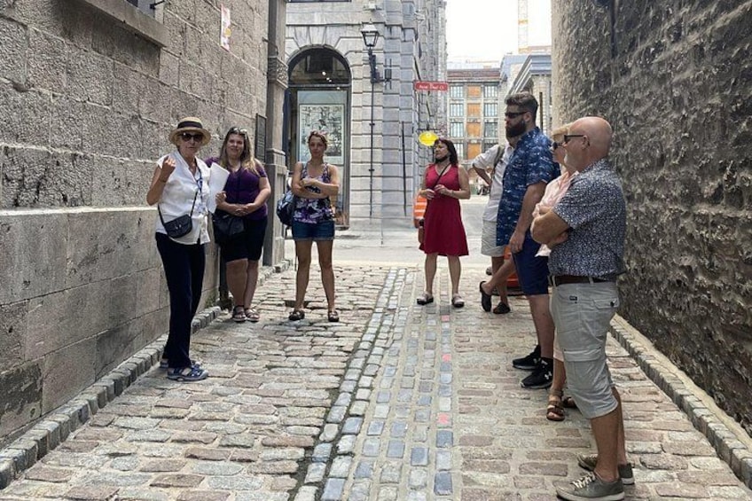 A group of people standing on a cobblestone street, a stone wall on the left, and a building with an arched entrance in the background. 2