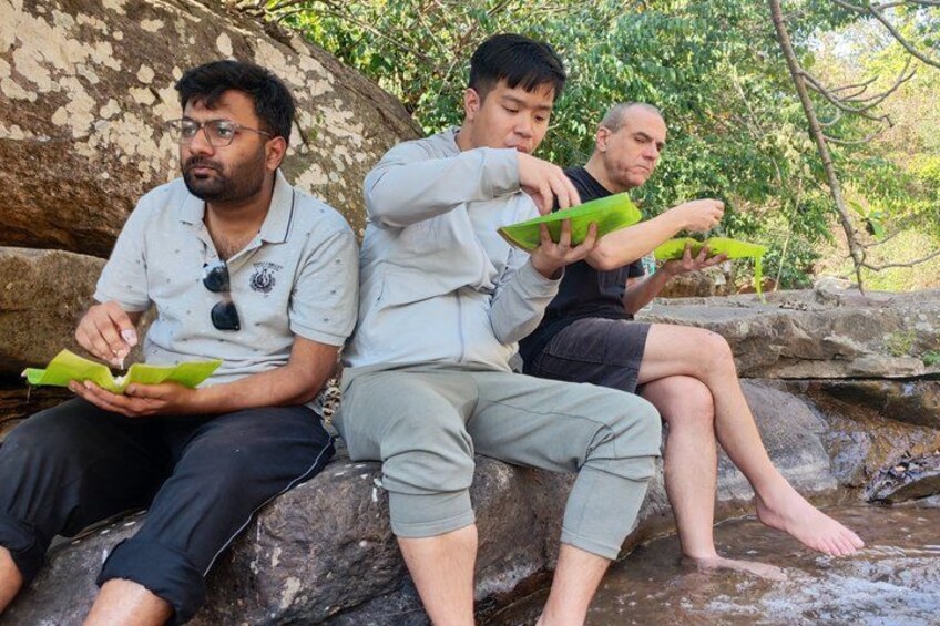 Tres personas sentadas sobre rocas junto a un arroyo, sosteniendo hojas verdes. 3