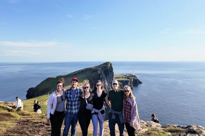 Un groupe de personnes se tient sur une falaise, surplombant une étendue d’eau, avec un phare en arrière-plan. 4