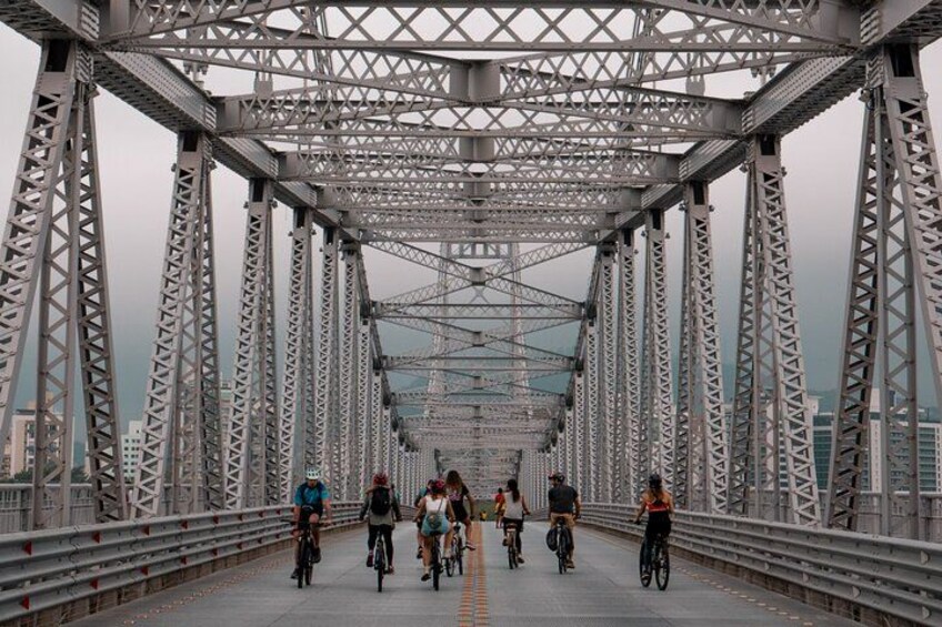 Un pont en acier à structure en treillis, des cyclistes sur la chaussée et des bâtiments de la ville en arrière-plan. 1