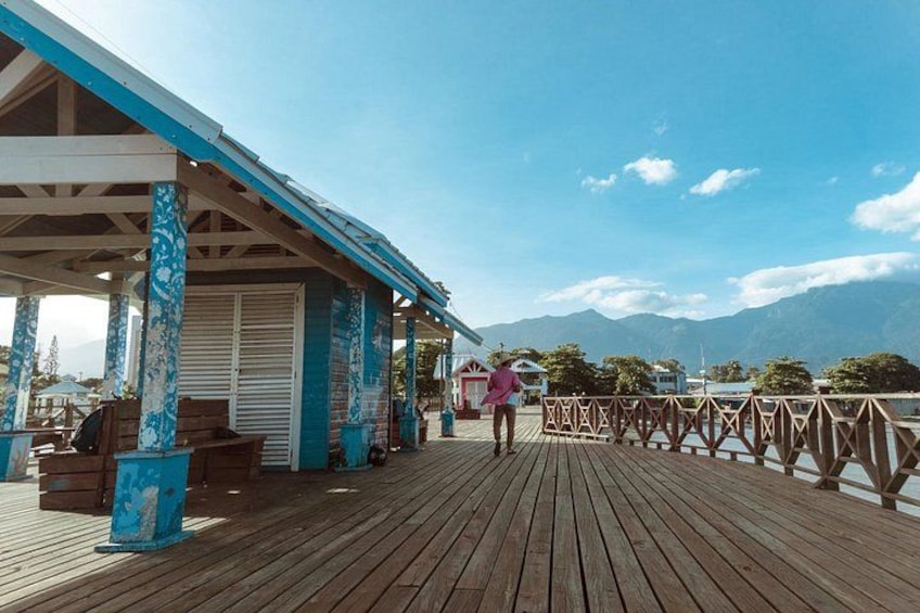 A wooden pier with a covered seating area, a person walking, and a mountainous landscape in the background. 1