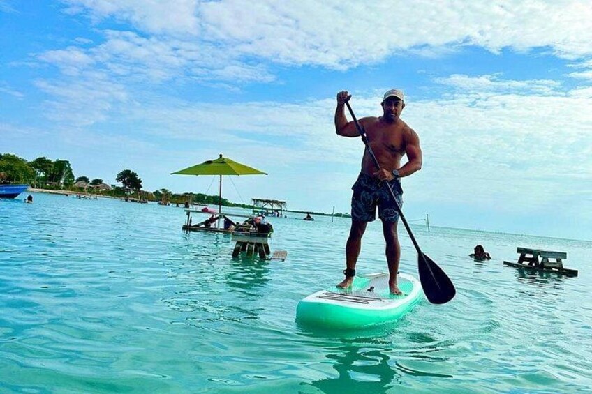 A man standing on a paddleboard in clear water, with a green umbrella and a picnic table in the background. 5