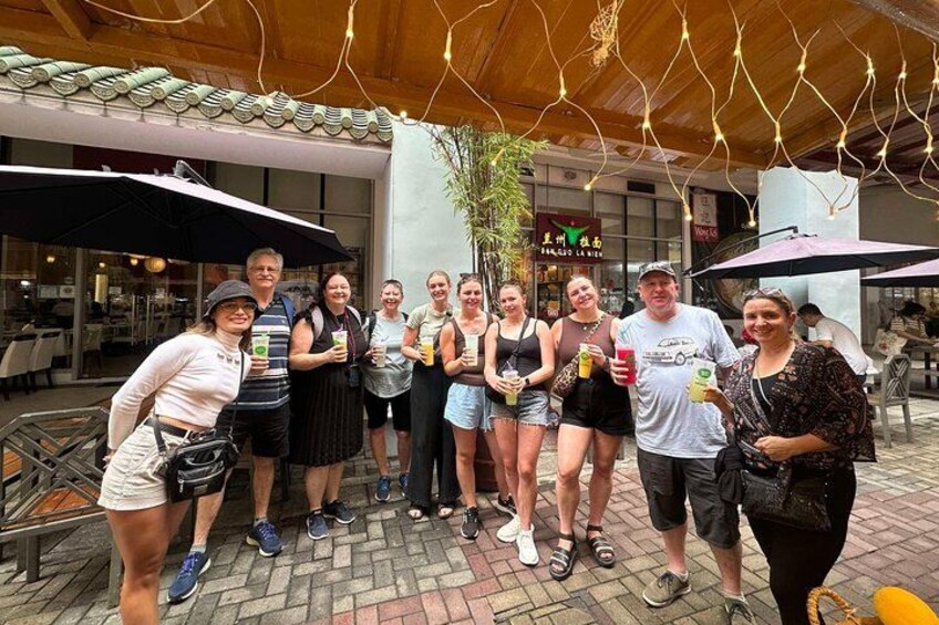 A group of people outdoors, some holding drinks, under a covered area with string lights. 3