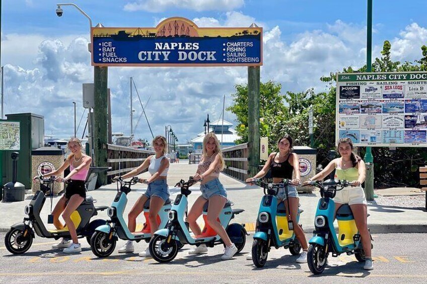 Four people riding electric scooters in front of Naples City Dock. 1