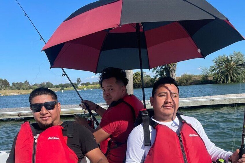Three people on a boat, wearing red life jackets, under a large umbrella. 3