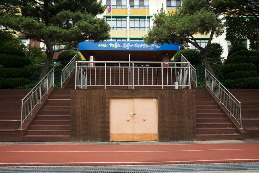 A set of stairs leading to a building with a blue awning, surrounded by trees and a red track. 3