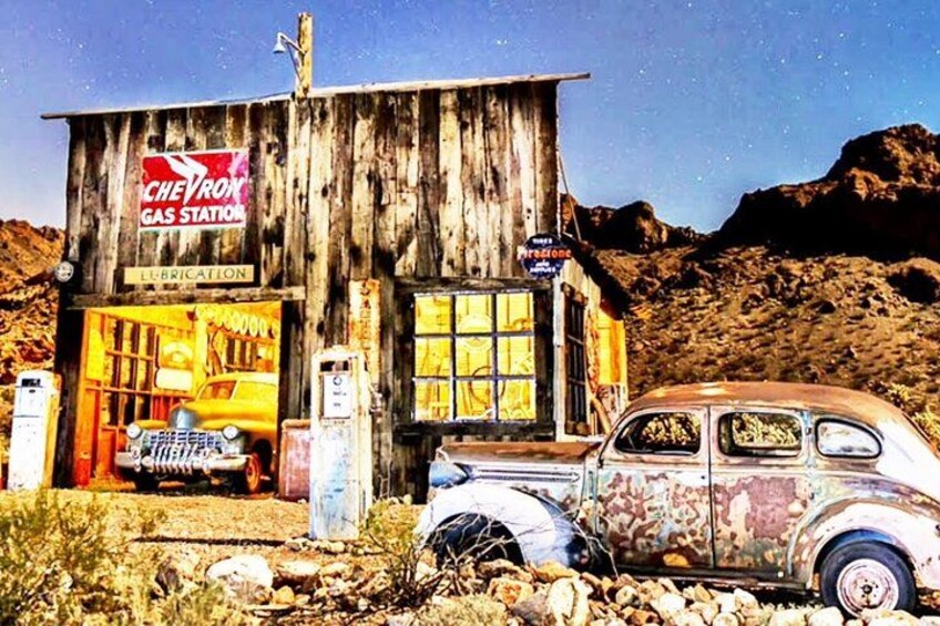 Une voiture ancienne est garée devant un bâtiment délabré portant l’inscription « STATION-SERVICE CHEVRON ». On aperçoit une pompe à essence et une voiture à l’arrière-plan. 1