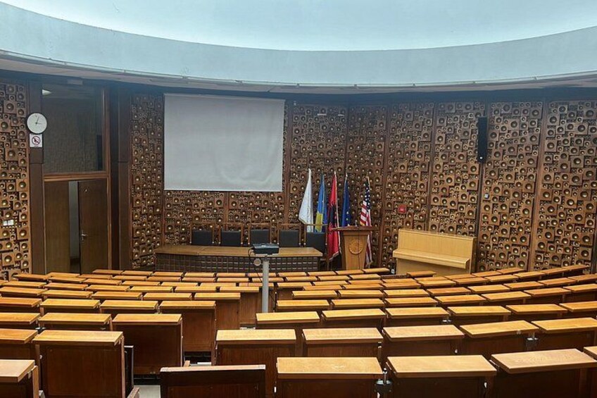 Un salón de conferencias con filas de sillas de madera, un atril y una pantalla grande. Banderas decoran la pared. 2