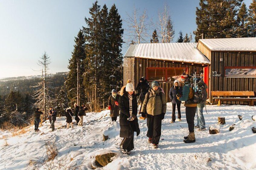 A group of people in winter clothing walking on a snowy path with a wooden cabin and trees in the background. 3