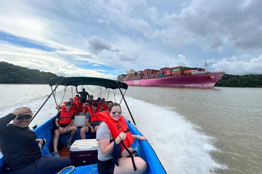 Un bote con pasajeros usando chalecos salvavidas, un barco de carga al fondo y una masa de agua con vegetación en la orilla. 3