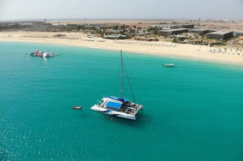 A coastal scene with a sailboat anchored near a sandy beach, a small boat in the water, and a distant shoreline with buildings and a clear sky. 2