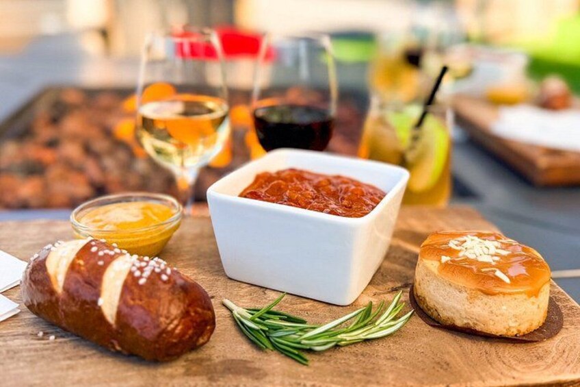 A wooden table with a pretzel, a bowl of tomato sauce, a small bowl of mustard, a bread roll, and a glass of beer. 1