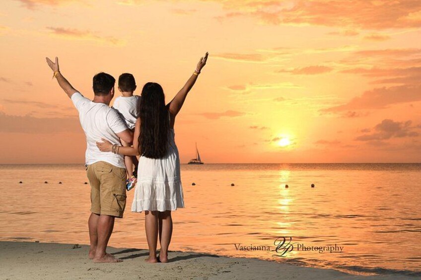 En familie på tre ved en strand i solnedgangen med oceanen og en sejlbåd i baggrunden. 1