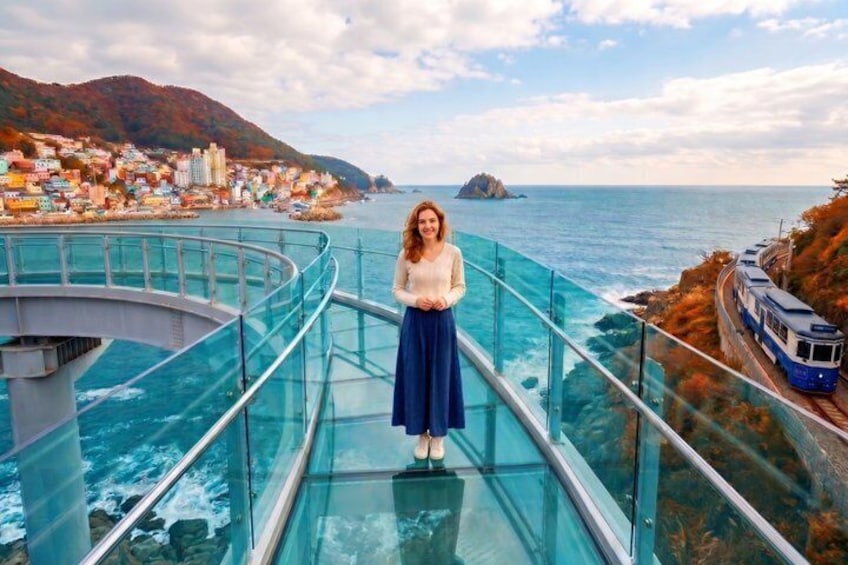 A person standing on a glass bridge over water, with a train on tracks to the right and a coastal town in the background. 1