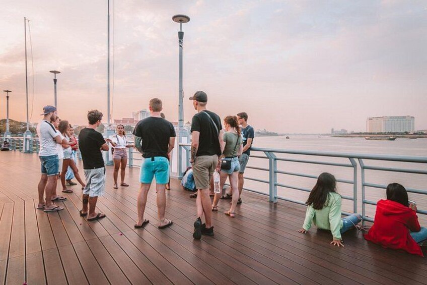A wooden pier with people standing and walking, overlooking a body of water with buildings in the distance. 4