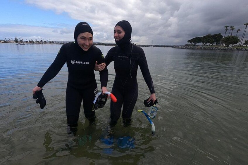 Two people in wetsuits standing in shallow water, holding diving equipment. 3