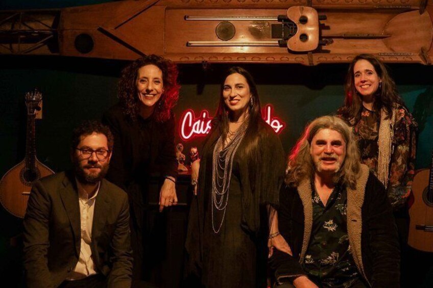 En gruppe mennesker i et svagt oplyst rum med guitarer og et neonskilt med teksten 'Cais'. 2