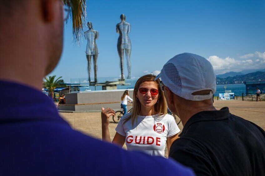 Une personne portant un t-shirt blanc sur lequel est imprimé le mot « GUIDE », se tenant devant une sculpture représentant une figure humaine. 3