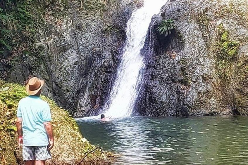 A waterfall, a person standing on a rock, and another person swimming in the water. 1