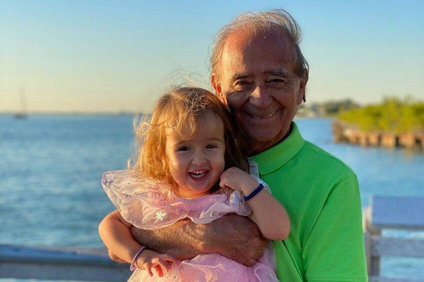 Une personne portant une chemise verte tient un enfant vêtu d’une robe rose, près d’un quai. 4