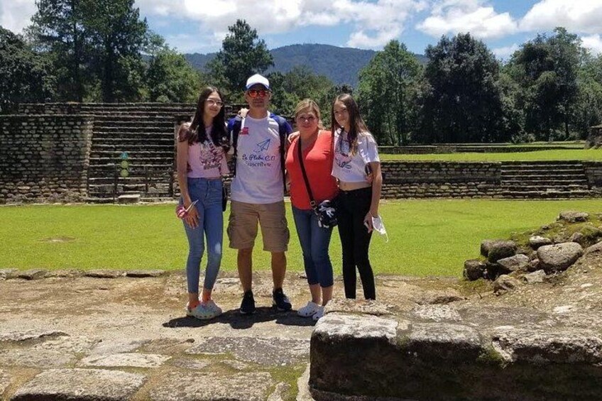 A group of people standing in an archaeological site with stone structures and a grassy field in the background. 2