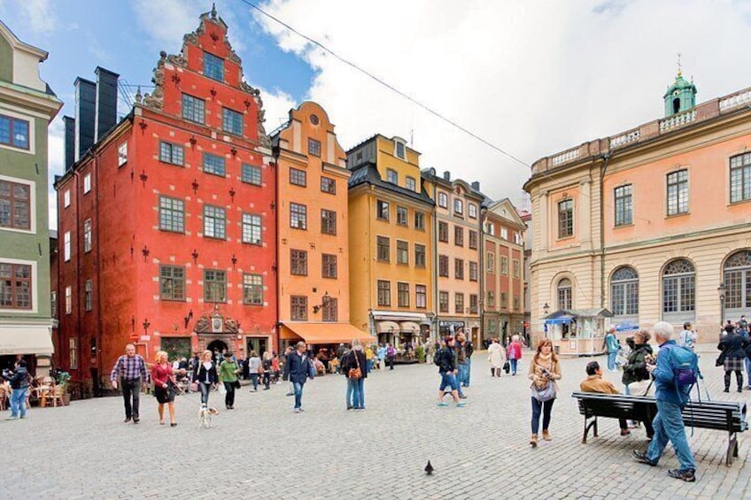 A cobblestone square with historic buildings, a fountain, and people walking. 1