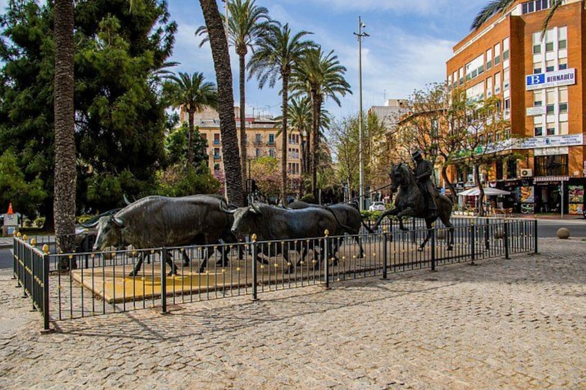 Une statue représentant une calèche tirée par deux chevaux, entourée d’une clôture, dans un environnement urbain avec des palmiers et des bâtiments en arrière-plan. 4