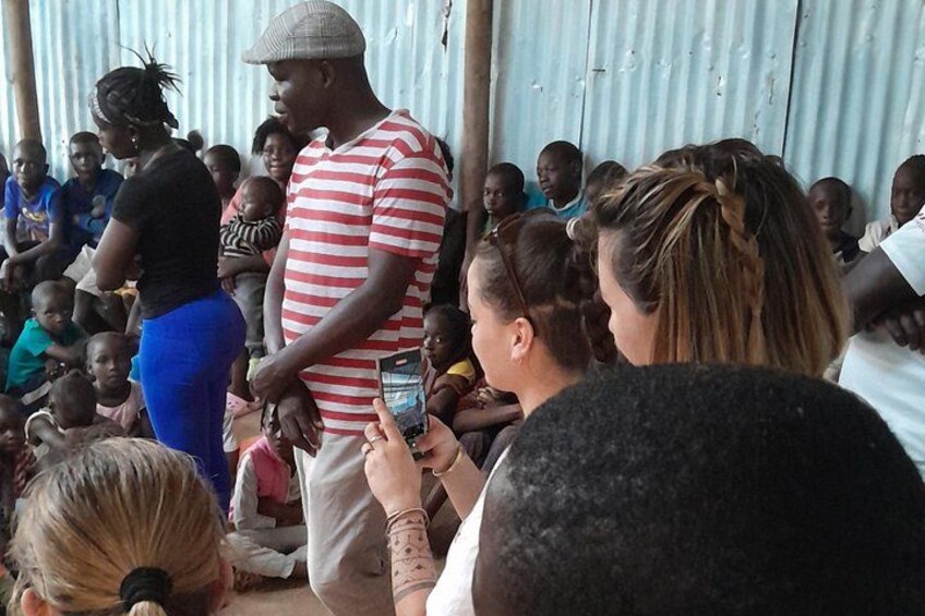A group of people gathered in an outdoor setting, some standing and some sitting, with a corrugated metal wall in the background. 4