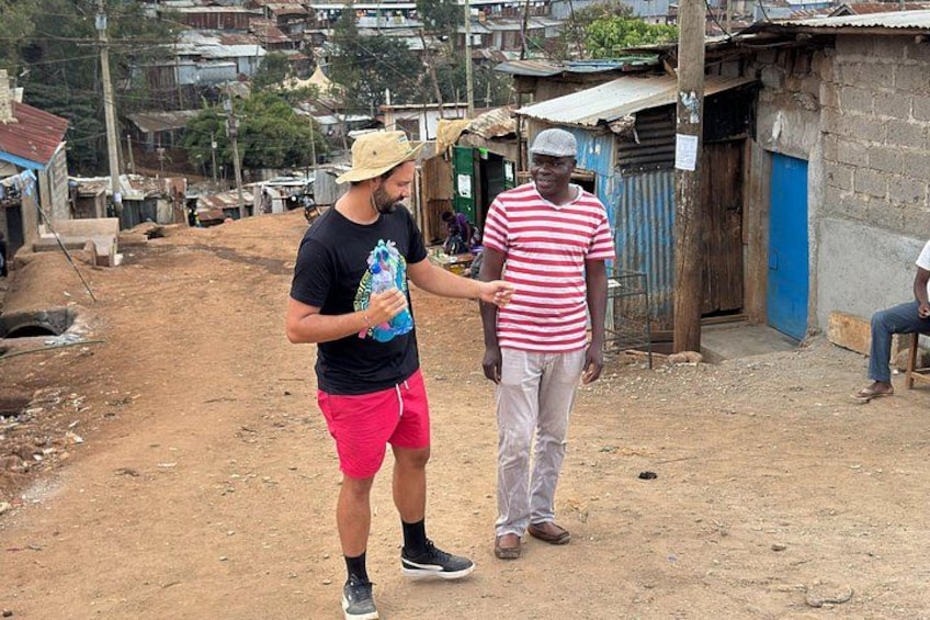 Two men standing on a dirt road in a rural area with makeshift buildings in the background. 1