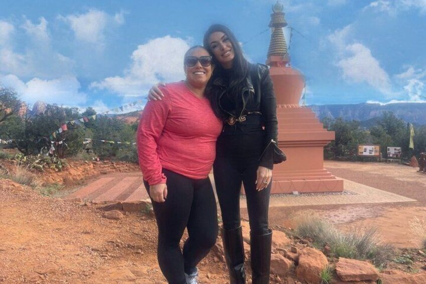 Two people standing in front of a red pagoda-like structure with a mountainous backdrop. 4
