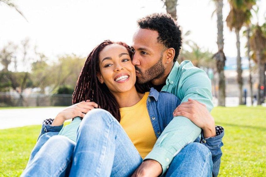Two people sitting on grass in a park with trees and a pathway in the background. 3