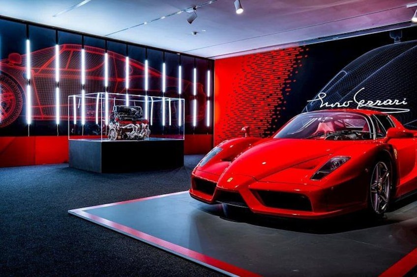 Un deportivo rojo en un concesionario con una decoración en tonos rojo y negro, un escaparate con un modelo de coche y una gran pantalla digital al fondo. 5