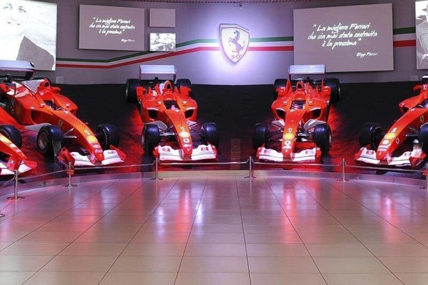 Tres autos de Fórmula 1 rojos exhibidos, con el logotipo de Ferrari y una cita en la pared. 4