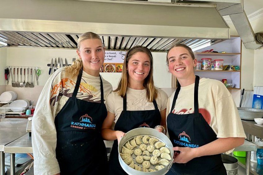 Three people wearing aprons with 'Kathmandu Cooking Academy' text, standing in a kitchen with utensils and a bowl of cookies. 1