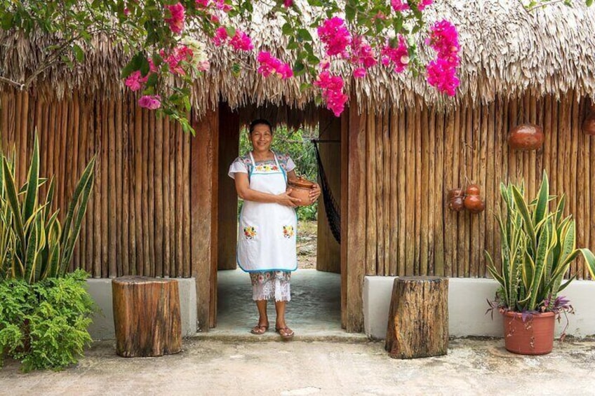 Una persona de pie frente a una cabaña con techo de paja, flores rosas y plantas en macetas. 1