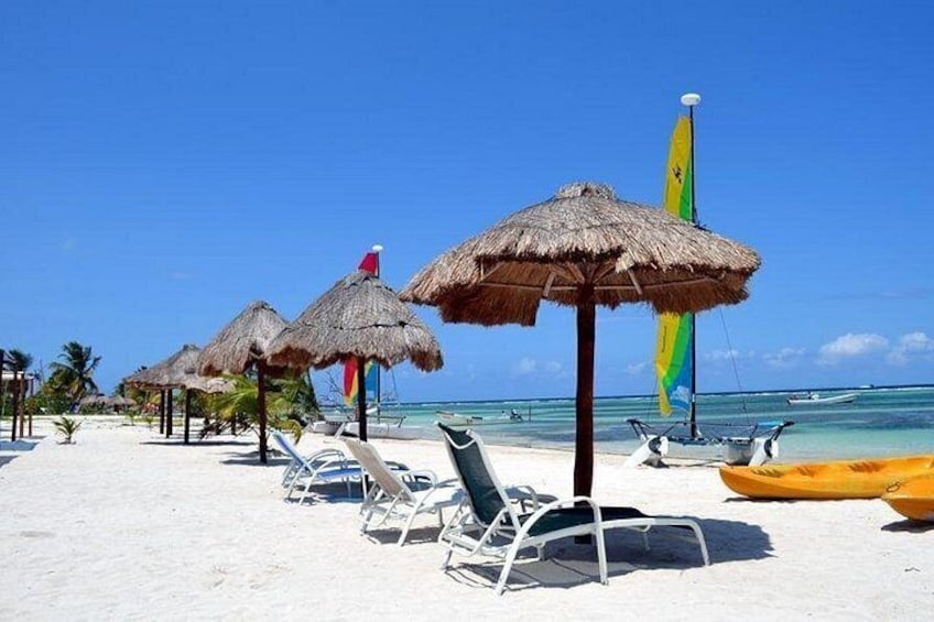 Una playa con sombrillas de paja, sillas de descanso y un velero. 3