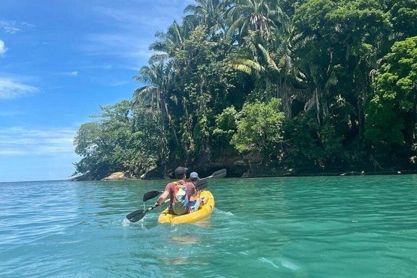 A person kayaking on a yellow raft in clear turquoise water, surrounded by lush green tropical vegetation and a rocky coastline. 1