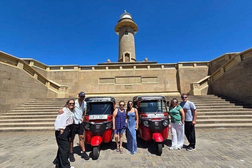 A group of people posing in front of a historic building with a tower, a red vehicle, and a blue vehicle. 1