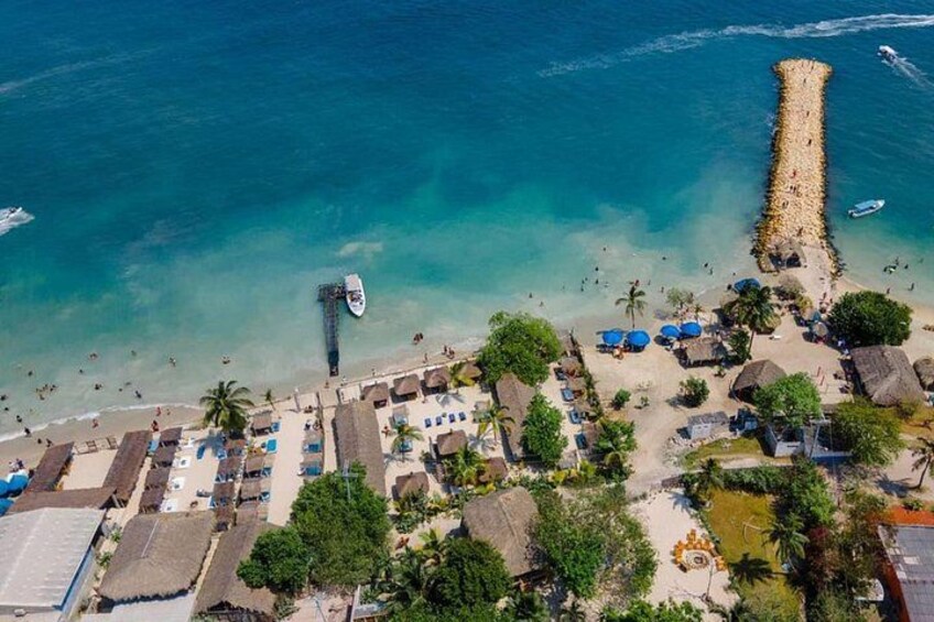 Luchtbeeld van een strand met helder turquoise water, een steiger en een klein bootje afgemeerd dichtbij de kust. 1