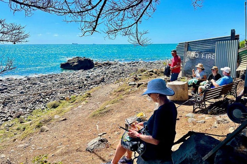 Una spiaggia rocciosa con persone sedute su panchine, un cartello con scritto 'NO FIRE' e una persona che usa un dispositivo mobile. 4