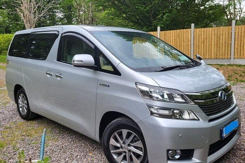 A silver minivan parked on a gravel driveway with a wooden fence and greenery in the background. 3
